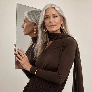 Woman with gray hair wearing a brown dress and gold jewelry, holding a mirror.